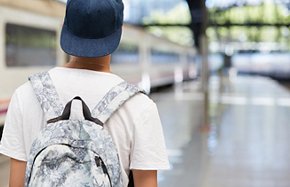 Back view of teenage school boy wearing white T-shirt and cap with a backpack on his back walking home from school. Urban background with copy space for your advertising content. Selective focus